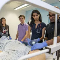 A group of students watch as one of them works on a dummy patient in a hospital bed