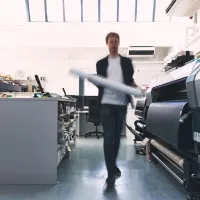 A student walking between several industrial-sized printers and a long work bench lined with guillotine paper cutters