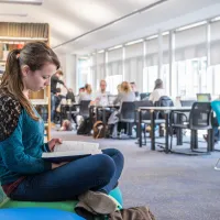 A student sits cross-legged reading a book in a bright, airy library. Behind her are shelves of books and other students working at desks.