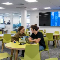 A wide shot of the software projects laboratory, with two students discussing a project at a table in the foreground, and more students working at computers and in a conference room further behind.