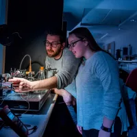 Two students set up a small experiment with a laser, in a darkened laboratory, surrounded by technical equipment.