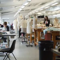 A wide shot of the fashion studio, full of students hard at work. To the right, a row of large workbenches are mixed in with over a dozen mannequins wearing various designs. To the left, a row of sewing machines.