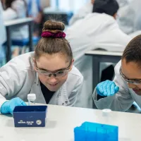 Two school children in lab coats and safety goggles examining laboratory equipment with focus, surrounded by others in a laboratory setting.