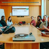  A group of nine people is seated around a wooden conference table, smiling and engaged in a meeting. Laptops are open, and a turntable with a vinyl record sits at the front of the table. A large screen behind them shows a web interface, and a whiteboard with notes is visible. The mood is friendly and collaborative in a modern, well-lit room.
