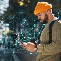 A man in a yellow beanie and olive-green sweater, engrossed in a smartphone, holding a disposable coffee cup, and wearing a backpack. He is standing against a backdrop of an abstract digital network design with interconnected nodes and lines.