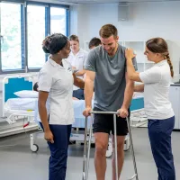 A patient, supported by 2 students, learns to walk using a walking frame.