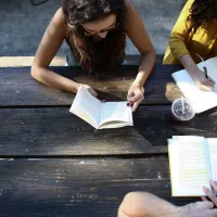 Three people gathered around at a picnic table while reading books and writing notes