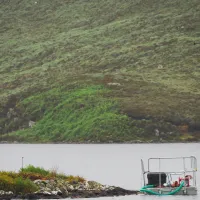 A drone hovers over a lake near a small floating platform with equipment, surrounded by lush green hills. The scene suggests a scientific or environmental study in a remote natural location.