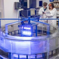 Students in white coats recording data from a rotating blue mechanical arm above a large circular transparent tank filled with liquid.