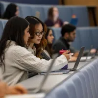 Two people sit side by side in a lecture theatre, looking at a laptop. One points at the screen while holding a cup. Other people sit in tiered seating behind, some using laptops.
