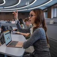 Accounting and taxation student sitting in a lecture with an open laptop on the desk. She has her hand raised to speak with the lecturer.