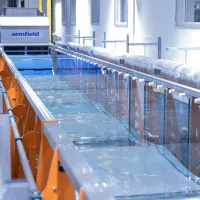A long water channel in a laboratory, featuring clear glass walls, orange triangular supports, and equipment along the sides, reflecting overhead lighting.