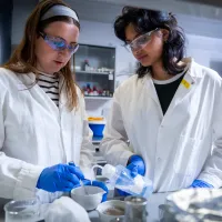Two students wearing lab coats, gloves and safety glasses preparing a laboratory sample.