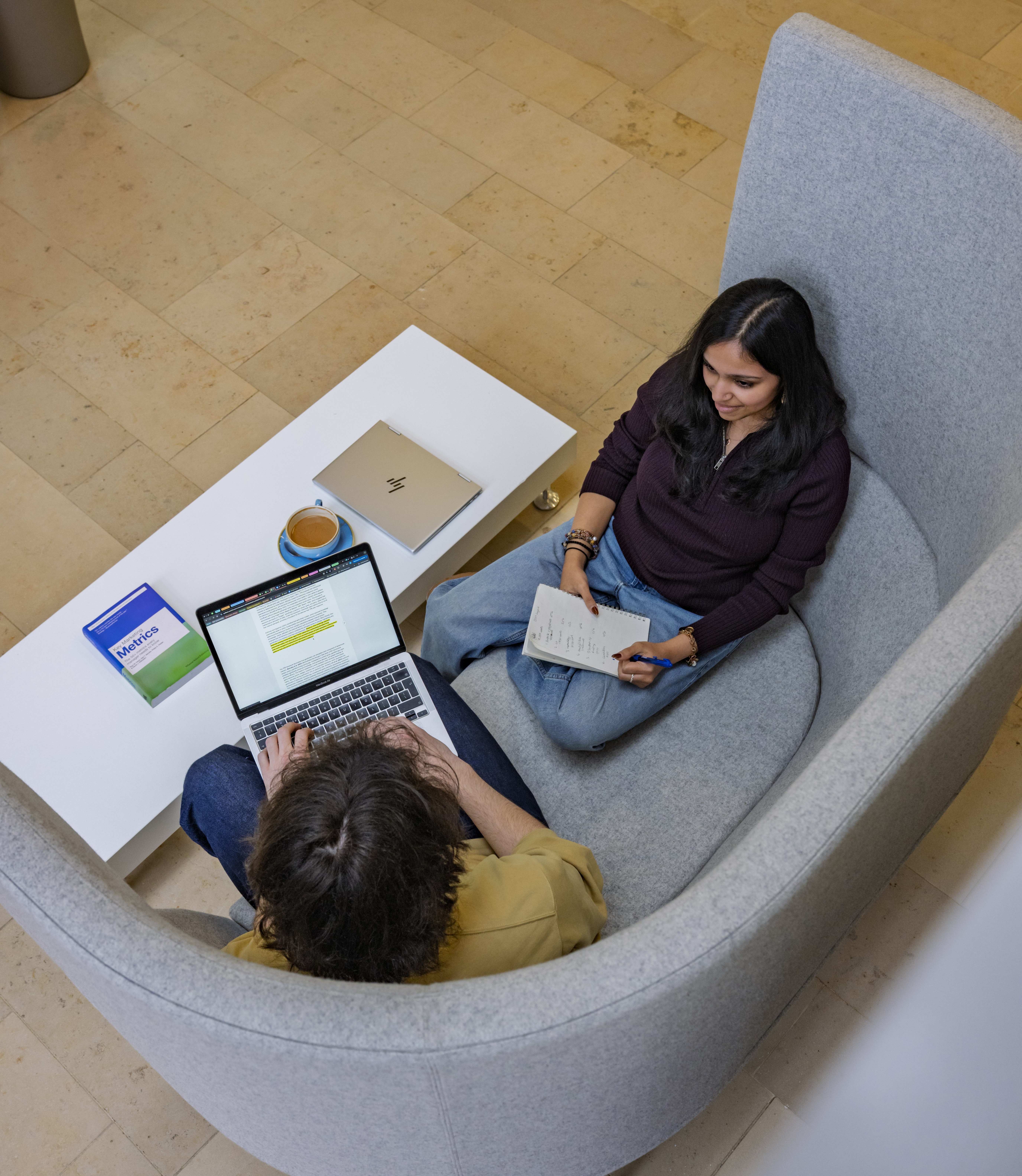 Looking down from a balcony at 2 students sitting on a sofa. They have laptops and books open on a table.