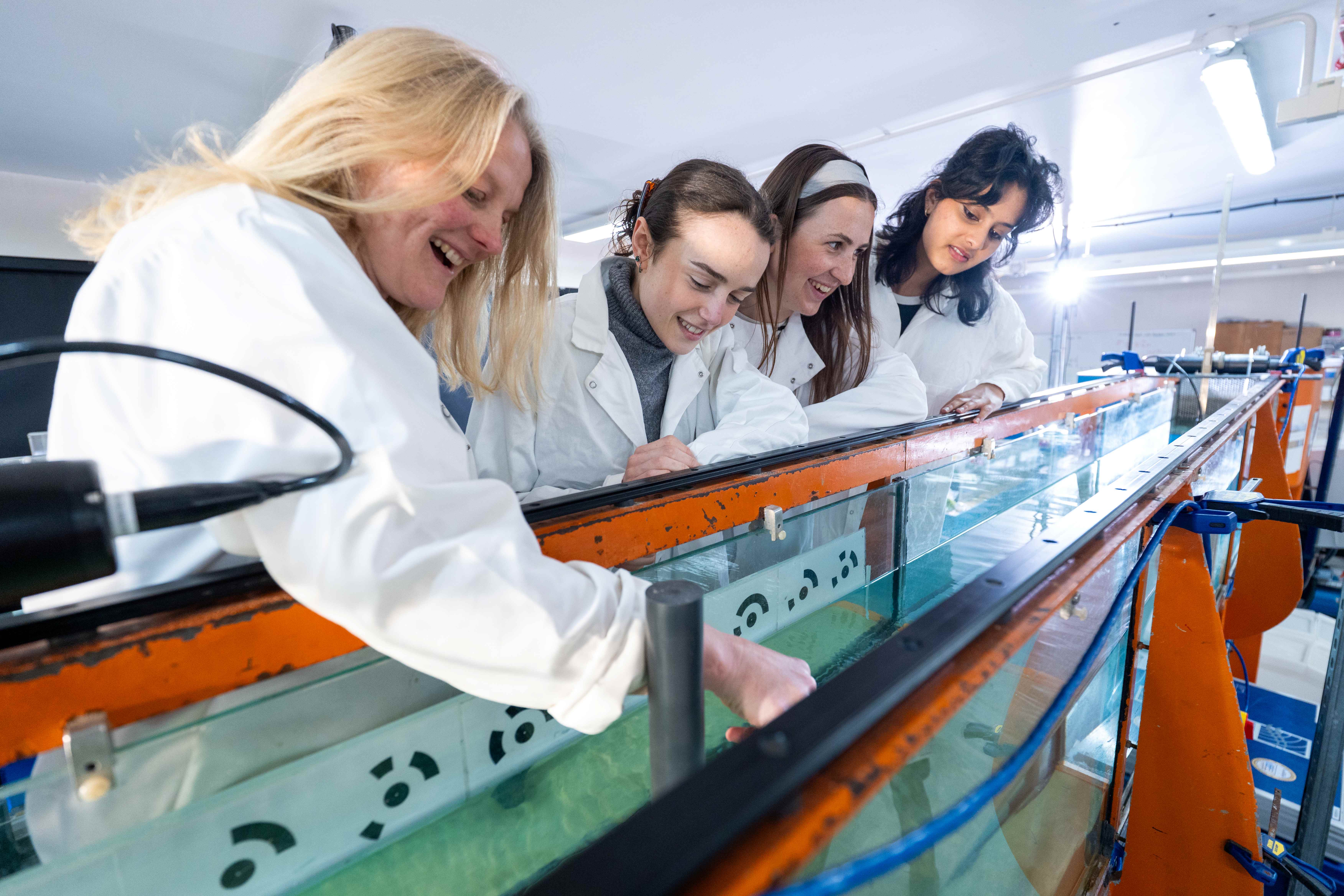 Four students in lab coats observing water flow in a laboratory flume tank.